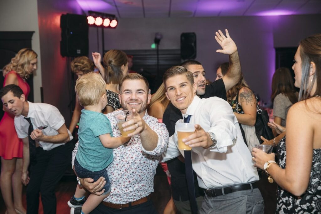 Two men dancing at wedding, smiling and pointing at the camera while one man holds a toddler.